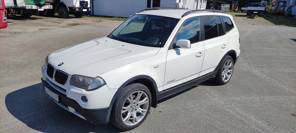 Biały SUV BMW X3 zaparkowany na asfaltowym placu, z widocznymi srebrnymi felgami, czarnymi zderzakami i charakterystyczną atrapą chłodnicy. W tle widać inne pojazdy i budynek.