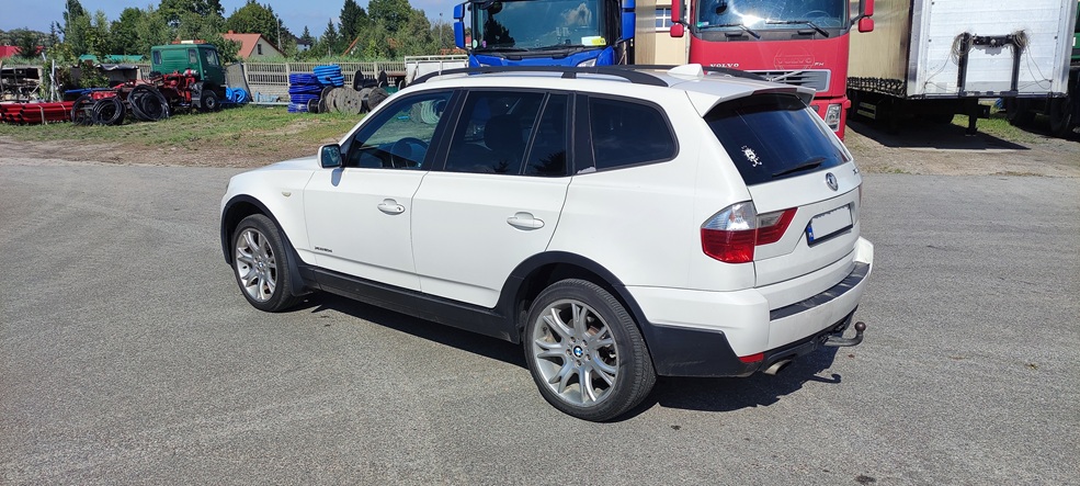 Biały SUV BMW X5 zaparkowany na asfaltowym placu, widok z tyłu i boku. W tle widać ciężarówki, maszyny rolnicze i ogrodzenie. Samochód ma czarne listwy nadkolenne, srebrne felgi i holownik na tylnym zderzaku.