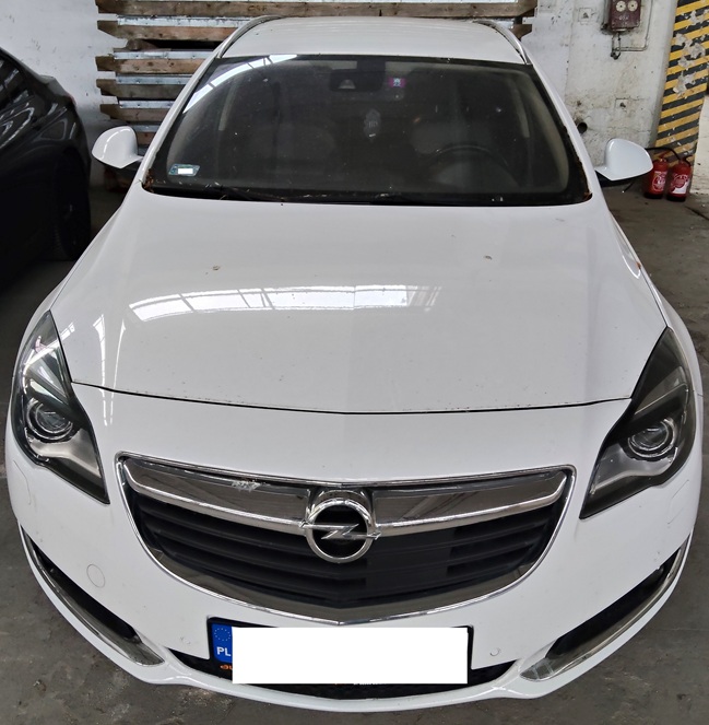 Biały samochód Opel z przodu, zaparkowany w garażu. Widoczne są reflektory, chromowana atrapa chłodnicy z logo Opel, tablica rejestracyjna z flagą Polski (zamazana), a w tle stosy palet i gaśnice.
