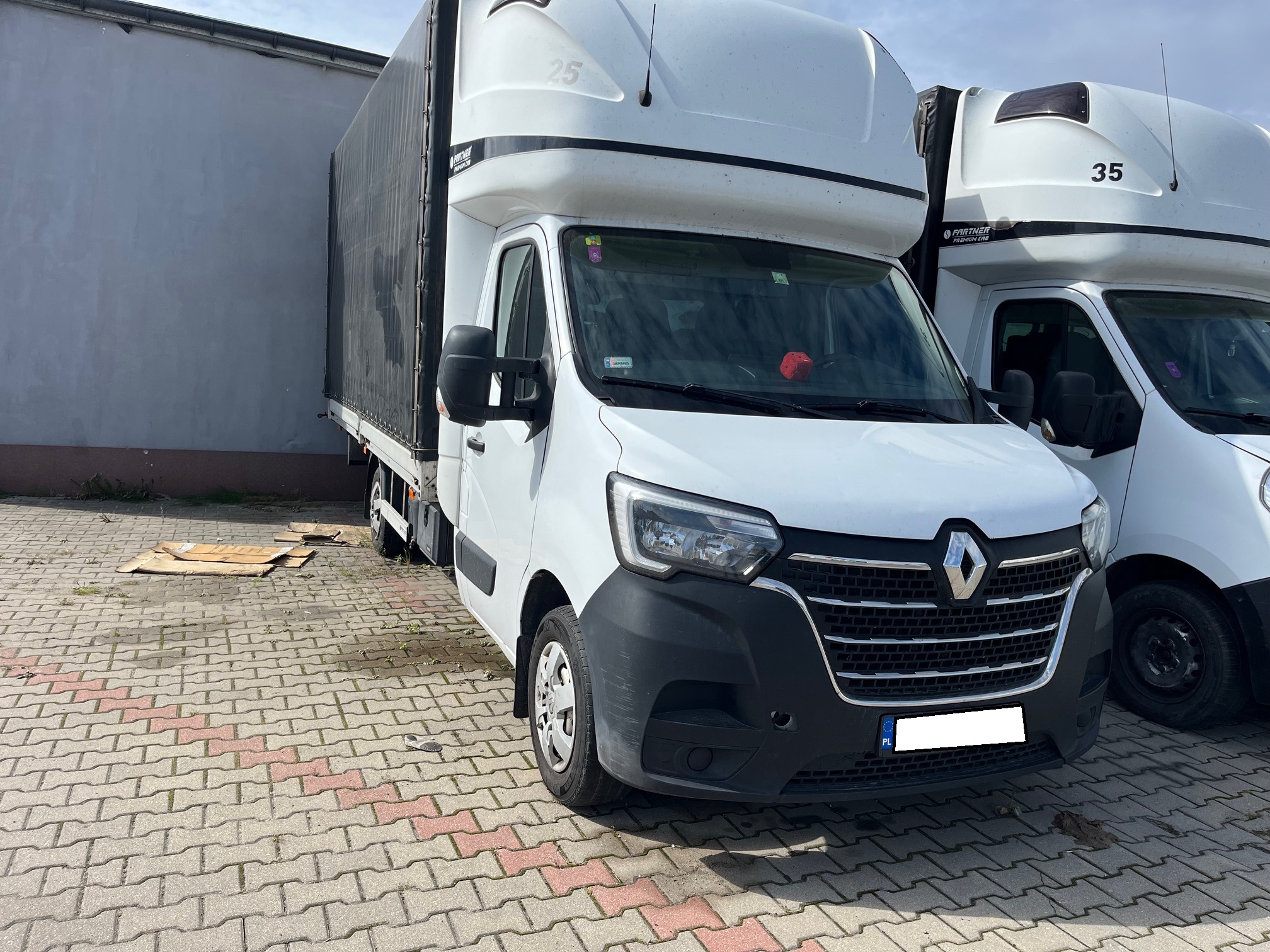 Biały furgon Renault z plandeką, oznaczony numerem 25, zaparkowany na brukowanym dziedzińcu obok innego podobnego pojazdu o numerze 35, przy szarej ścianie budynku.