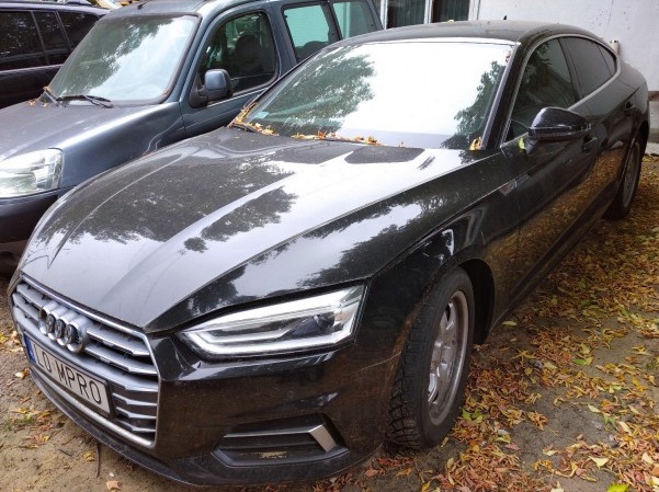 Czarny samochód Audi A5 zaparkowany na zewnątrz, otoczony suchymi liśćmi. Widoczny jest przód pojazdu z charakterystyczną atrapą chłodnicy i reflektorami. Obok stoi inny, ciemny samochód.