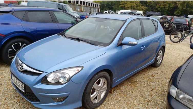 Jasnoniebieski samochód Hyundai i30 z tablicą rejestracyjną „OJ 8293E” stoi na żwirowym placu. W tle widoczne są inne pojazdy, w tym motocykl i rower, oraz budynek. Na szybie przedniej widoczny napis „33/25”.